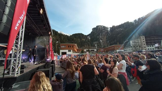 Konzertarena in Hintertux, links auf der Bühne steht Josh mit Band, rechts das . gefüllte Areal. Im Hintergrund die Talstation der Gletscherbahn, eine Gondelbahn fährt noch