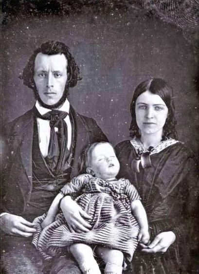 An antique black-and-white photo from the 1840s. There are three people: a father, a mother, and a baby. 

Father is rather tall and has a beard style which was fashionable at the time: the front of his face is clean shaven, and his beard wraps around his chin and into his sideburns. He is slightly balding at the top of his head, near the front of his hairline. His hair is a dark color.

The lady is smiling only slightly. She has dark-colored hair that ends in ringlet curls near her neck. 

In Mother’s arms is a sleeping baby, who seems to be about the right age to be taking her first steps and running around just a little. Baby is wearing a striped dress and is deeply asleep in her mother's arms. The baby's mouth is a bit agape, and her head is tilted back a bit. Her cheeks have just a bit of shading to them, which was probably rosey pink in real life.