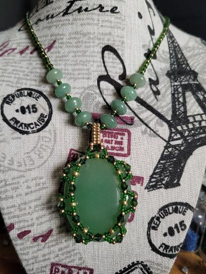A green aventurine thumb stone is put into a netted bezel comprising of 4mm bicones, 8/0 gold seed beads, and 11/0 translucent green seed beads. The pendant is strung on a coordinating necklace of mentioned seed beads and an set of 8x10mm green aventurine rondelles.