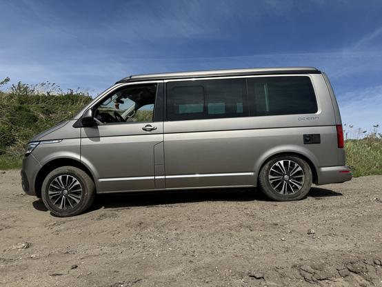 A Volkswagen California T6.1 in the sun on gravel, photographed from the side. The left front wheel is e tended quite far down (for a Transporter) while the rear left week is almost all the way up.