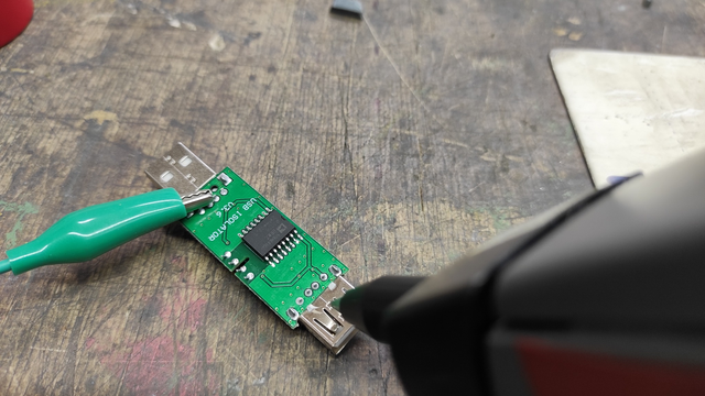 The mentioned 'USB isolator v3.6' on a wooden desk, with a green wire attached to one of the protruding pins on the USB-A plug, visible alongside the ESD gun tip applied to the socket side.