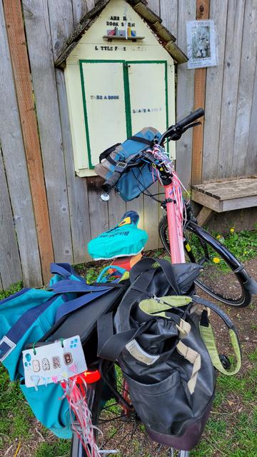 Photo of a salmon colored bike with a turquoise bag on the left side of the back rack and a black bag on the right side. The seat is covered by a turquoise seat cover. A light blue handlebar bag hangs from the front. The bike is parked in front of a wood fence with a little free library in the shape of a house hung on the fence. At the top of the house wooden letters spell out Barbara's Book Barn. On the doors letters spell out take a book leave a book. A few letters are missing but you can still see their silhouettes to read the words.