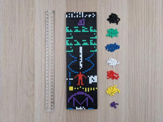 A photo of a 3D printed mosaic representing the Arecibo Message, a 23x73 graphic transmitted into outer space in 1974. It's made up of multiple different colours, which are shown in small piles on the right. On the left is a 30cm ruler for scale.