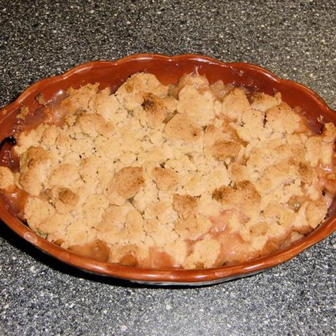 Eine ovale, braune Backform steht auf einer dunklen Arbeitsplatte. Die Form enthält einen gebackenen Crumble mit goldbrauner Krümeldecke. Unter den Krümeln scheint eine fruchtige Füllung hervor, die an den Rändern karamellisiert ist. Farblich lässt sich ein Kontrast zwischen der dunklen Oberfläche und dem hellen Crumble erkennen. Die Textur der Krümel ist ungleichmäßig, was auf eine hausgemachte Zubereitung hindeutet.