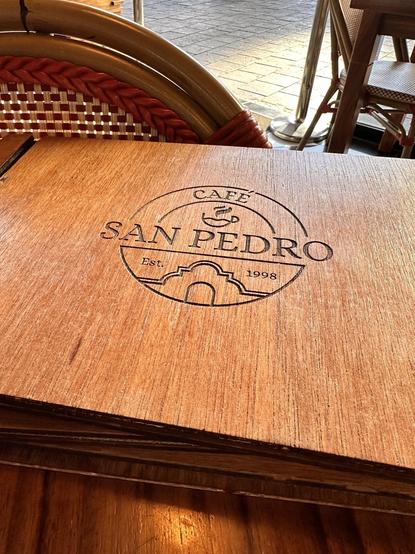 Wooden café table with the logo "Café San Pedro" engraved on it, and a wicker chair in the background.