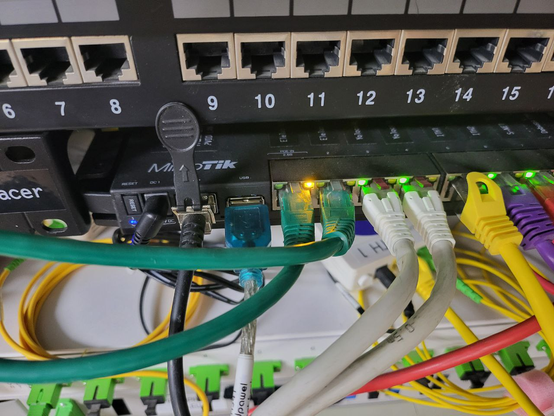In the photo, a patch panel with sockets labeled from 6 to 15 is visible. Below it is the Mikrotik RB5009UG+S+, with variously colored Ethernet cables, one SFP, and a power cable plugged into it, along with a USB cable. The continuation of this cable is described in the post and visible in subsequent photos. Below the Mikrotik, a fiber optic patch panel is visible.