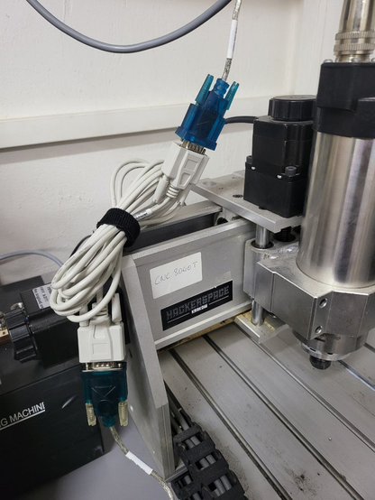 The photo shows the other end of the cable plugged into the USB port of the Mikrotik - a transparent blue RS232 connector on a silver-shiny cable plugged into one end of a null-modem cable terminated with female RS232 connectors on both sides. The null-modem cable is fastened with Velcro as its full length is not needed, so it can be coiled up to serve as an adapter. Also visible in the photo is a mini CNC milling machine, the 3040T, located under the hackerspace network cabinet.