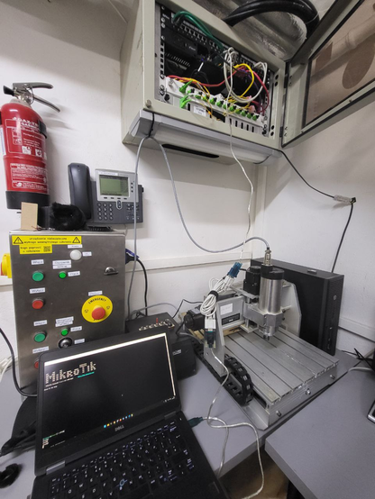 The wide-angle photo captures the entire scene, showcasing an open network cabinet containing the aforementioned Ethernet patch panel, Mikrotik router, fiber media converter, and fiber patch panel. A cable hangs from the cabinet, connected via a null-modem cable to a laptop placed below. On the laptop's monitor, the Mikrotik terminal is visible, displaying the characteristic "MikroTik" ASCII MOTD from the MikroTik RouterOS. Additionally, the photo shows a CNC 3040T milling machine and its controlling computer, spindle controller, and a large metal control cabinet for the CNC with various red and green buttons and indicators. In the middle of the cabinet is a yellow-red mushroom emergency stop button. Each button and indicator is labeled with a sticker, while yellow stickers on top of the control cabinet indicate required training. Above the power supply cabinet, a fire extinguisher and a Cisco 79xx series phone, part of the Hackerspace's VoIP installation, are mounted on the wall.