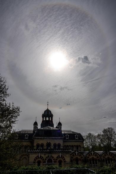 A halo surrounds the sun in a sky with many light wispy clouds, below is an ornate Victorian Building with several towers