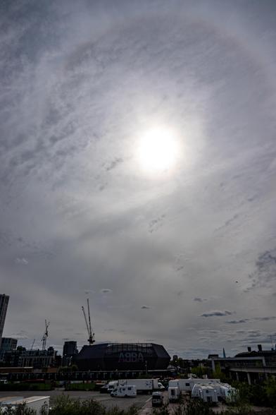 A half-halo is barely visible around the top of the sun, in a sky with many light wispy clouds. Below is a large building with "ABBA" written on it.