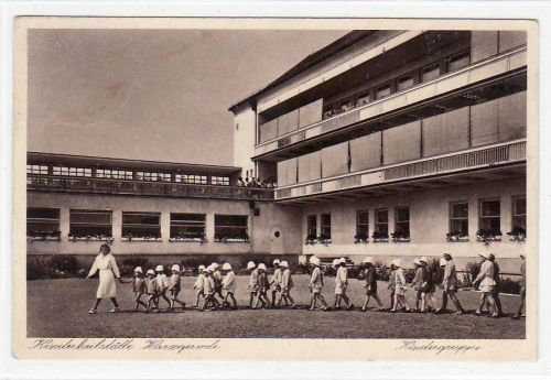 Historische Postkarte. Schwarz weiß Fotografie der ehemaligen Klinik (im Hintergrund) mit einer Gruppe Kinder im Vordergrund, angeführt von einer Schwester im weißen Kleid. Die Kinder gehen in Zweierreihen und tragen Sonnenhüte.