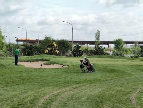 Ein Mann mit grünem Shirt steht auf einem Golfplatz in einem Sandbunker. Mitte Bild vor dem Grün, auf dem die Fahne steckt, steht sein Golftrolley , mit diversen Golfschlägern. Im Hintergrund sieht man eine Brücke stehen, die aber keinen Bezug zum Golfplatz hat