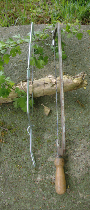Two hack saws with their blades stuck into a thick but crumpling branch lied on a mossy stone. The blades look straight. The left one is made of wire, and the right one out of thick metal and has a wooden handle.