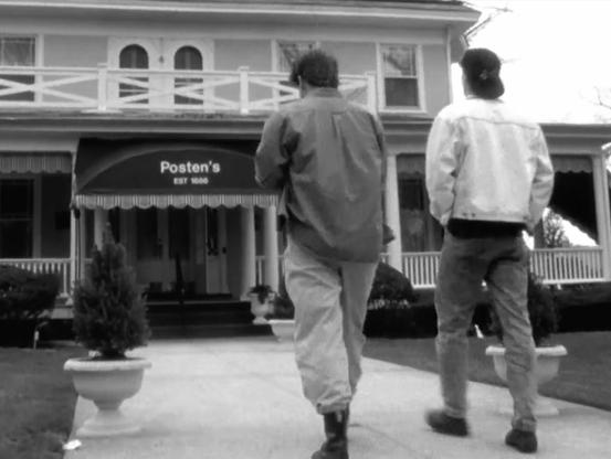 A black and white screencap from a film of two young men walking towards a funeral home.