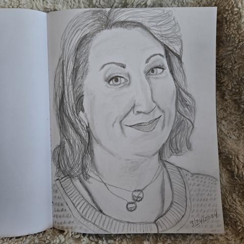A sketchbook is open to a pencil sketch of Maggie Service, playing Maggie in Good Omens, Season 2. Her hair is parted at the side and brushed off her face, falling in soft waves at the bottom. She has her head turned to the right and smiles slightly at the viewer. She wears a white t-shirt under a cardigan sweater and two round pendants on delicate chains around her neck.