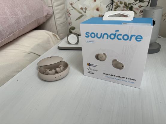 Anker Soundcore Sleep A20 mit Ladecase und Verpackung auf einem Nachttisch.
