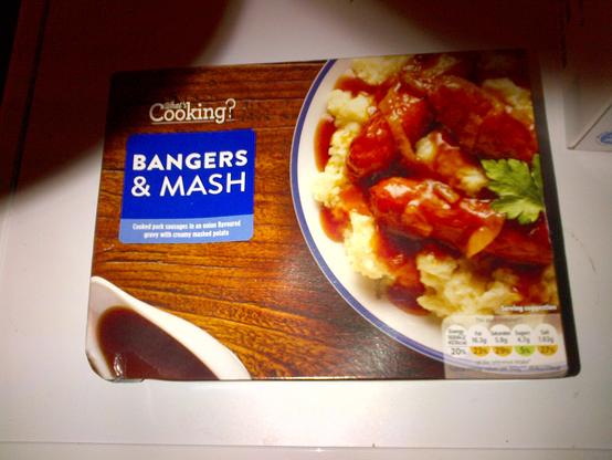 A box of "What's Cooking?" brand bangers and mash ready meal from Lidl, with a write on blue brand label and a picture of a plate of bangers and mash and a gravy boat.  The box is sitting on an imperfectly flashlit white countertop.