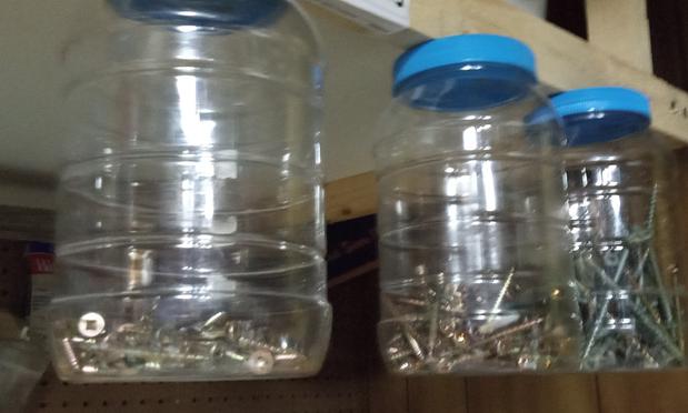 Three clear plastic jars attached to the underside of a shelf in the workshop. Each size of screw length is in a different jar