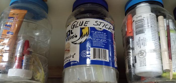 Plastic mayonnaise (clear) jars affixed below a shelf. Filled with glue sticks, markers, glue tubes, ect