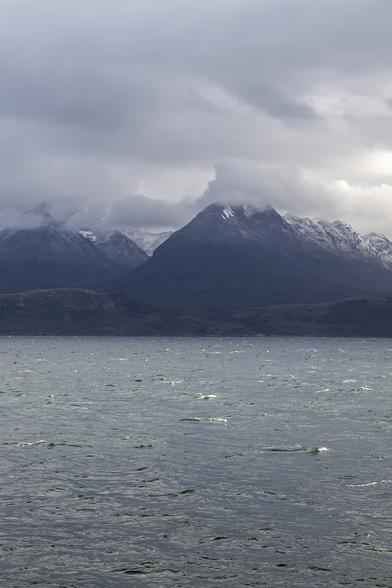 A portrait view taken from the water and looking back towards land. It's a very overcast day with layers of grey cloud overlapping one another above. The sea is choppy and feels very cold and uninviting. The landscape beyond the water is dark and menacing too with barely any distance from the shore to where mountains with jagged peaks rise, the nearest of them topped with cloud that hides their uppermost points, further parts of these steep ranges visible in the distance, topped with snow broken up by black crevasses. The scenery is jaw-droppingly wonderful but you wouldn't want to be here without many layers of clothing.