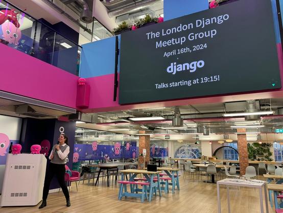 Cagil Ulusahin Sonmez, one of the organizers of the Django London Meetup, is pictured on her feet, microphone in hand, doing the introductory speech to kick off the evening. The backdrop is a large presentation screen that reads 'The London Django Meetup Group, April 16th, 2024, Talks starting at 19:15!' with the Django logo. The event is hosted in a vibrant and modern office space with playful octopus-themed decor.