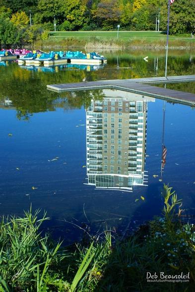 Photograph of a building reflected in a clear pond with paddle boats and an American flag, surrounded by lush greenery. Image at:  https://beautifulsunphotography.com/featured/urban-mirage-deb-beausoleil.html See more art & blog at: https://beautifulsunphotography.com/ https://debbeautifulsunphotography.com/ https://www.zazzle.com/store/beautifulsun_designs https://debbeausoleil.com