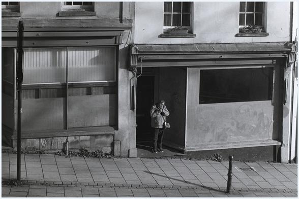 A black and photograph of a woman on St Michael's Hill, Bristol, UK who has interrupted her progress to step into a vacant shop doorway, out of the morning wind and sun, in order to light her cigarette.