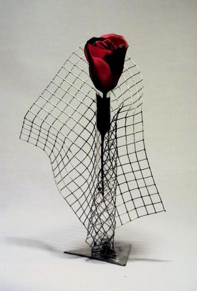 Eine Rose in einem Reagenzglas ist in ein verknittertes Gitter aus verzinktem Draht eingewickelt, das Gitter ist auf ein dreieckiges Edelstahblech als Fuß geschraubt.
A rose in a test tube is wrapped in a crumpled grid of galvanised wire, the grid is screwed onto a triangular stainless steel sheet as a base.