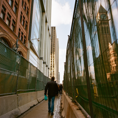 Midday pedestrians walk a narrow path through a large downtown construction project, concrete bollards and high fences guide them. The left side, above the fencing, tall buildings loom and, on the right, the clock tower of Toronto’s Old City Hall is a shadow through the scrim on the fence.