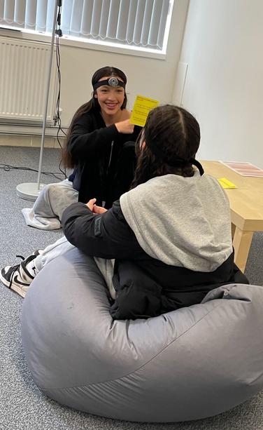 Two teenagers sitting on beanbags face each other. Both are wearing head cameras. The camera is visible as a small grey plastic circle attached to a headband, which goes around the forehead. One teenager is holding a card up and is smiling. The other has their back to the camera and their face is not visible.