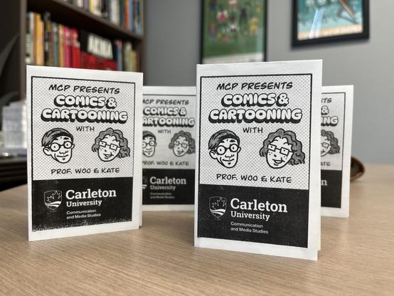 Several copies of a small black and white zine, stood up on a table. The cover of the zine has two cartoon heads and reads, “MCP Presents Comics & Cartooning with Prof. Woo & Kate.”