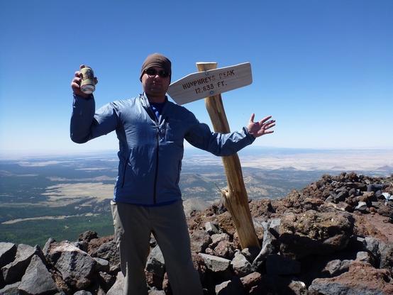 A person stands next to a signpost marking "Humphreys Peak 12,633 FT." atop a rocky summit, holding up a can, with a clear blue sky and vast landscape in the background.