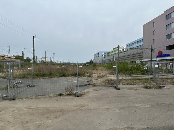 Blick auf ein mit Baustellenzaun umzäuntes Brachland. Links am Bildrand ein Bahndamm, rechts ein langezogenes, rechteckiges, graues Büro-, Supermarkt- und Parkgebäude.