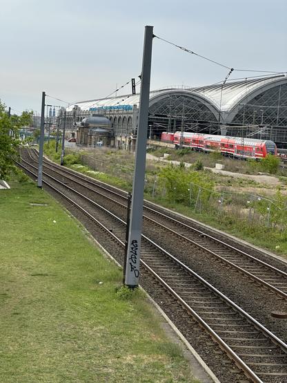 Blick auf den Hauptbahnhof Dresden mit gewölbter Decke und Glasfront. Ein roter Doppelstockzug verlässt den Bahnhof.
Auf der linken Bildseite führen Straßenbahnschienen entlang.
Zwischen Bahnhof und Straßenbahnschienen ein verwaistes, mit Pflanzen überwuchertes Bauland.