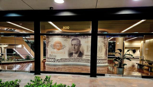 View into clear windows of a Federal Reserve Bank on Water Street, Downtown, Jacksonville.  The lobby glows goldish-yellow from interior lighting where a very large imitation copy of the US one-hundred thousand dollar bill, featuring President Woodrow Wilson is on display.