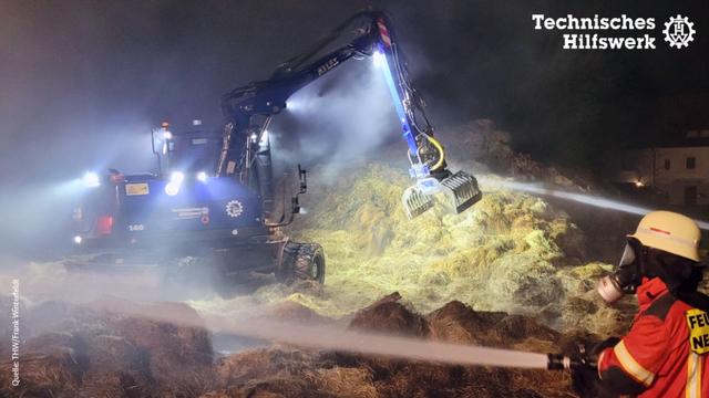 Ein Bagger steht inmitten von qualmenden Stroh- und Heuballen. Im Vordergrund ist ein Mensch in Feuerwehr-Einsatzkleidung zu sehen. Er trägt eine Atemschutzmaske.