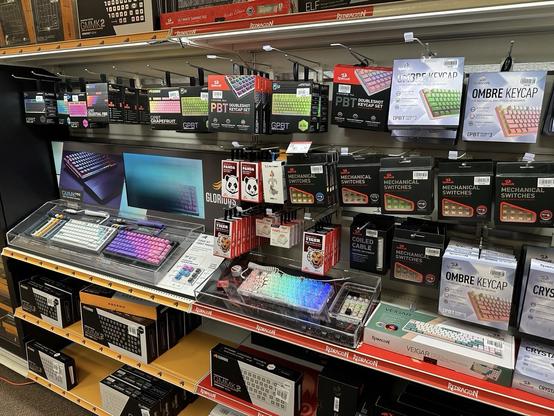 A selection of computer keyboard accessories on retail display shelves, including keycap sets, mechanical switches, and coiled cables.