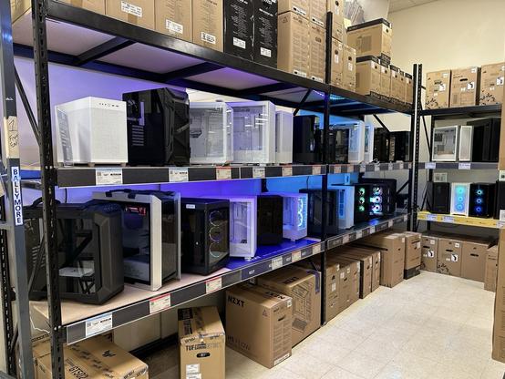 A variety of computer cases displayed on metal shelving in a store, with some featuring LED lights. Boxes are stacked on the bottom shelves and floor.