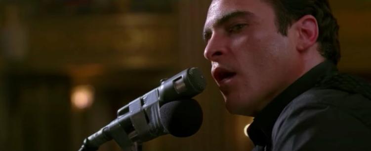 A close shot of a man on a stage, singing into the mic, looking over at someone standing next to him