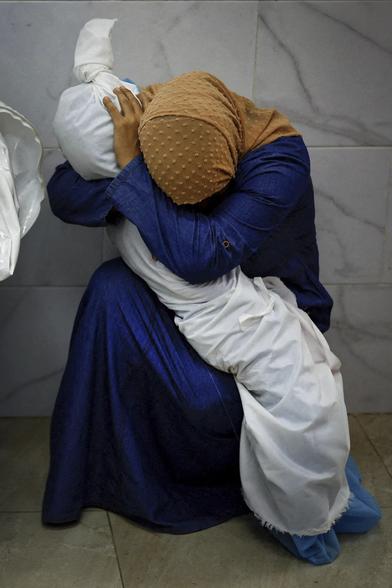 Photography of Palestinian woman dressed with a single blue outfit bending her head covered by an orange-brown piece of material cradling the body of a dead kid all wrapped in a single white sheet she is holding the body both firmly and tenderly as if she wouldn't want to let it go her right hand bent elbow surrounding the dead kid's neck  the palm of her left hand is pressing the wrapped head against women's right elbow thus the head seems locked like in a soft wrestling catch