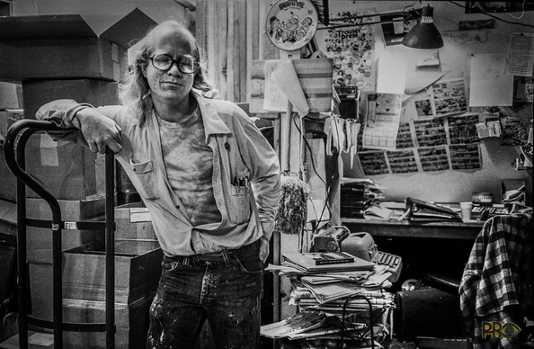 A black and white photo of a man leaning against a warehouse trolley. He has glasses, long hair - but almost nothing on his skull, an open shirt all the way down to his belt and a T-shirt underneath. Many pens in the breast pocket, paint stains on dark trousers. Behind him a stack of cardboard boxes to the left and to the right an office workplace overflowing with stacks of letters and a jumble up the wall.

 🇸🇯Et sort hvit foto av en kar som lener seg mot en lagertralle. Han har briller, langt hår – men nesten ingenting på skallen, åpen skjorte helt ned mot beltet og T-skjorte under. Mange penner i brystlomma, malingsflekker på mørk bukse. Bak ham en stabel pappesker til venstre og til høyre en kontorarbeidsplass overfylt med brevbunker og et virvar oppetter veggen.