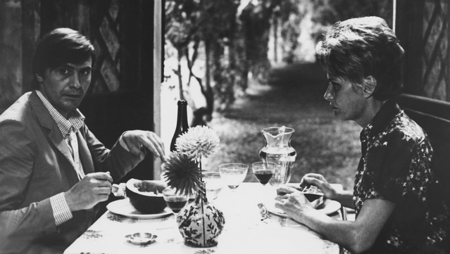 Athos Magnani (Giulio Brogi) and Draifa (Alida Valli) sharing a meal. There's a carafe with water, a small vase with two flowers, glasses of wine, and half a melon. Athos looks up, past us, towards something.