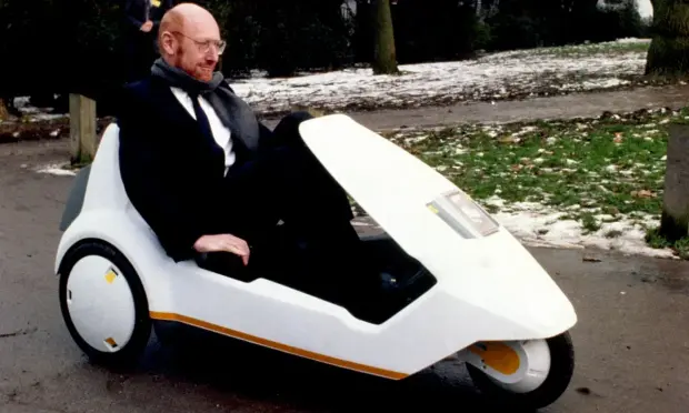 Sir Clive Sinclair riding in a Sinclair C5. It's a single person three-wheeled vehicle, best described as somewhere between a bike and a car. The design has the sleek dazzling veneer of the 1980s, in pure white with a yellow-brown accent stripe.