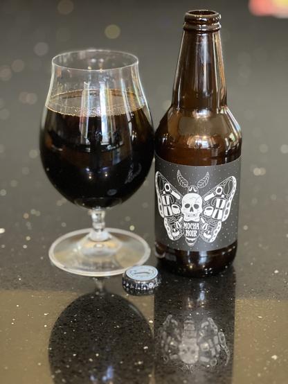 A tulip glass of dark dark beer. The 16 ounce bottle is to the right with a black label and a white stylized butterfly with a skull as its thorax