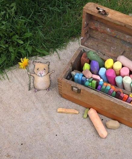 Streetart. A small hamster with a flower has been painted with chalk on a beige sidewalk next to a meadow. He stands proudly next to artist David Zinn's paint box, which is full of colorful pieces of chalk. In his hand he is holding a yellow flower with a long stem. Title: "Saul has appointed himself Chief Dandelion Officer and Herald of the Chalkbox."