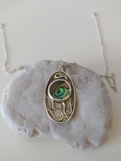 a pendant placed on a pebble. it's oval shaped with a green round gemstone and it has oxidized patterns