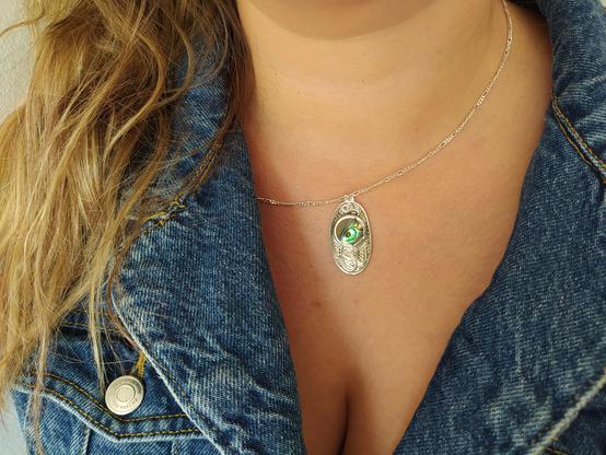 an oval shaped pendant with a green round gemstone and it has oxidized patterns. it's worn by a woman with denim shirt