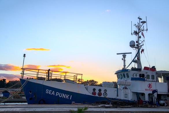 das rettungsschiff "sea punk one" am pier von der seite fotografiert mit gegenlicht vom sonnenunterhgang, welches den himmel blau-golden färbt