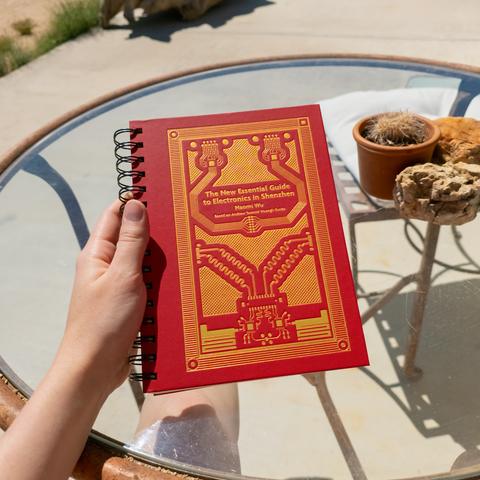 Photo of Lety sitting at a glass table on a concrete slab in the desert on a bright sunny day. Anything she is wearing is not visible as the photo was taken from her POV. In the foreground, Lety is holding “The New Essential Guide to Electronics in Shenzhen” by Naomi Wu in her left hand above the glass tabletop, the subtitle on the cover reading “Based on Andrew ‘bunnie’ Huang's Guide”. The spiralbound book is closed, and its red cover with embossed gold circuit board design is displayed. In the background, a rock and a small pot containing a cactus are on the glass table, and Lety's bare left leg is visible under the table's clear surface. She feels the need to clarify that she is not at home and that the little cactus in the pot is not her's because the little cactus in the pot is very dead.