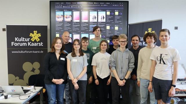 Die Hacker des Hackathons vom Kultur Forum Kaarst. Im Hintergrund ein Bildschirm mit Veranstaltungen des Kino Kaarst und GBG.